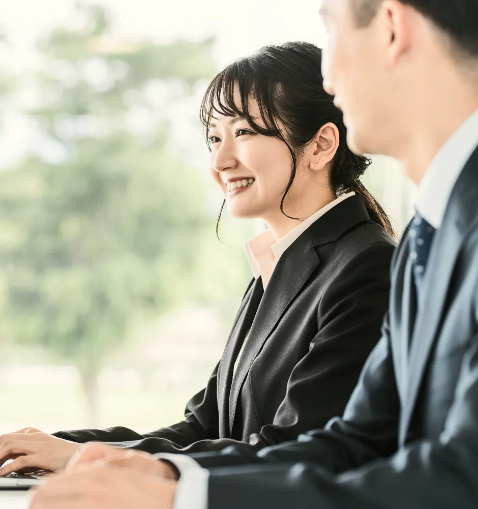 明るいオフィスでスーツ姿の女性が微笑みながらパソコン作業をする様子、隣には同僚の男性が座っているビジネスシーン